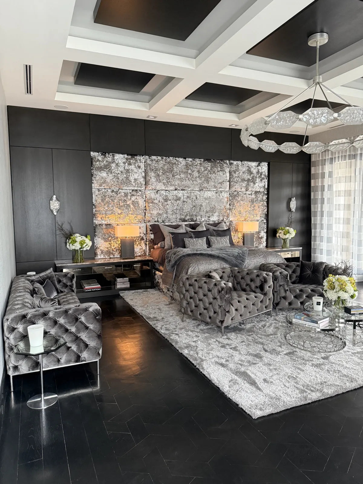 Black bedroom seen from the seating area, the distressed silver upholstered headboard wall lit from within between dark joinery