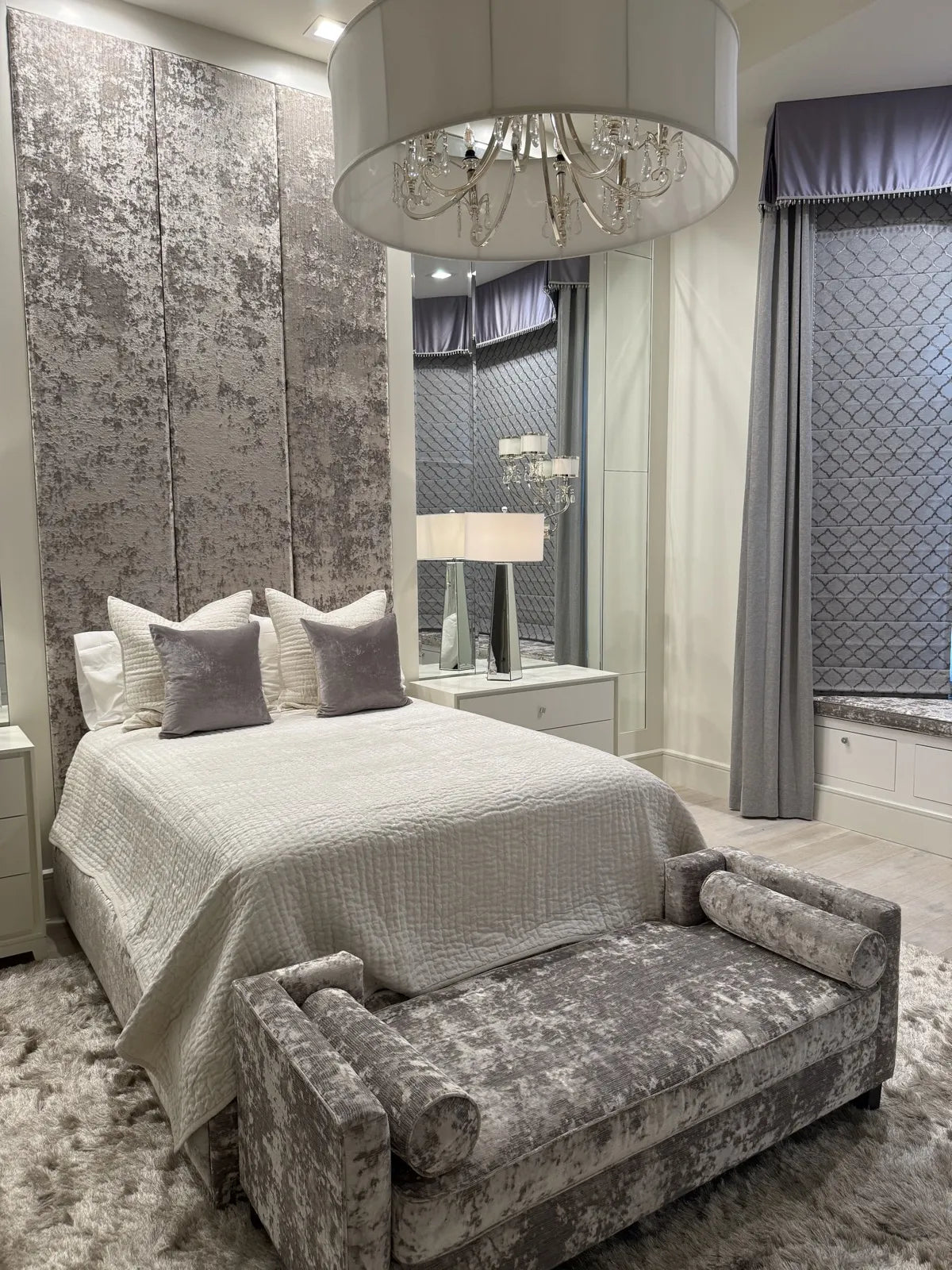 Bedroom with a full-height upholstered headboard wall in crushed silver velvet, flanked by mirrored panels and a mirrored nightstand
