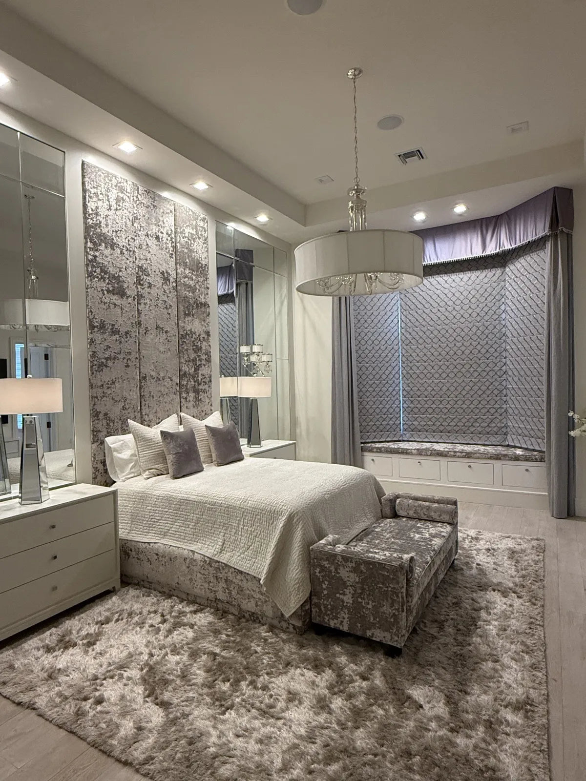 Wide view of the same bedroom showing the upholstered headboard wall, the bay window seat and a matching crushed velvet bench at the foot of the bed