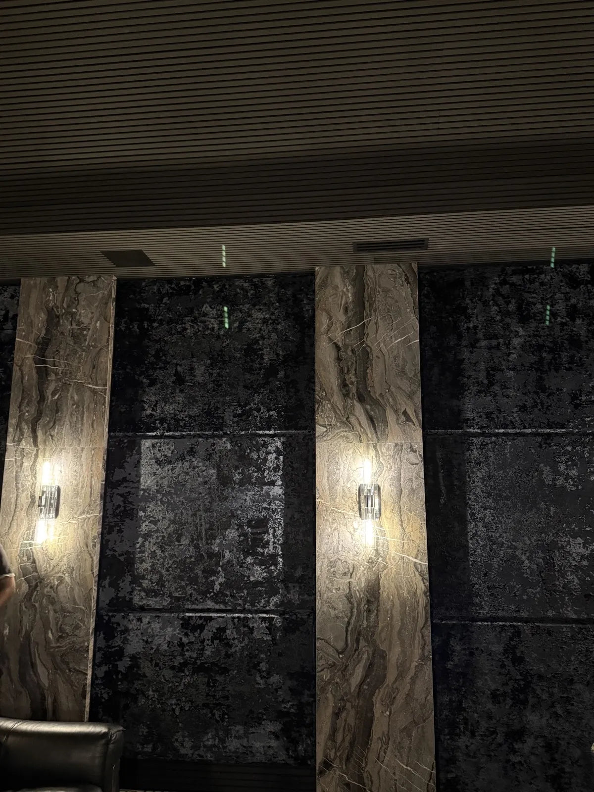 Black distressed upholstered wall panels divided by marble pilasters with wall sconces, in a home theater