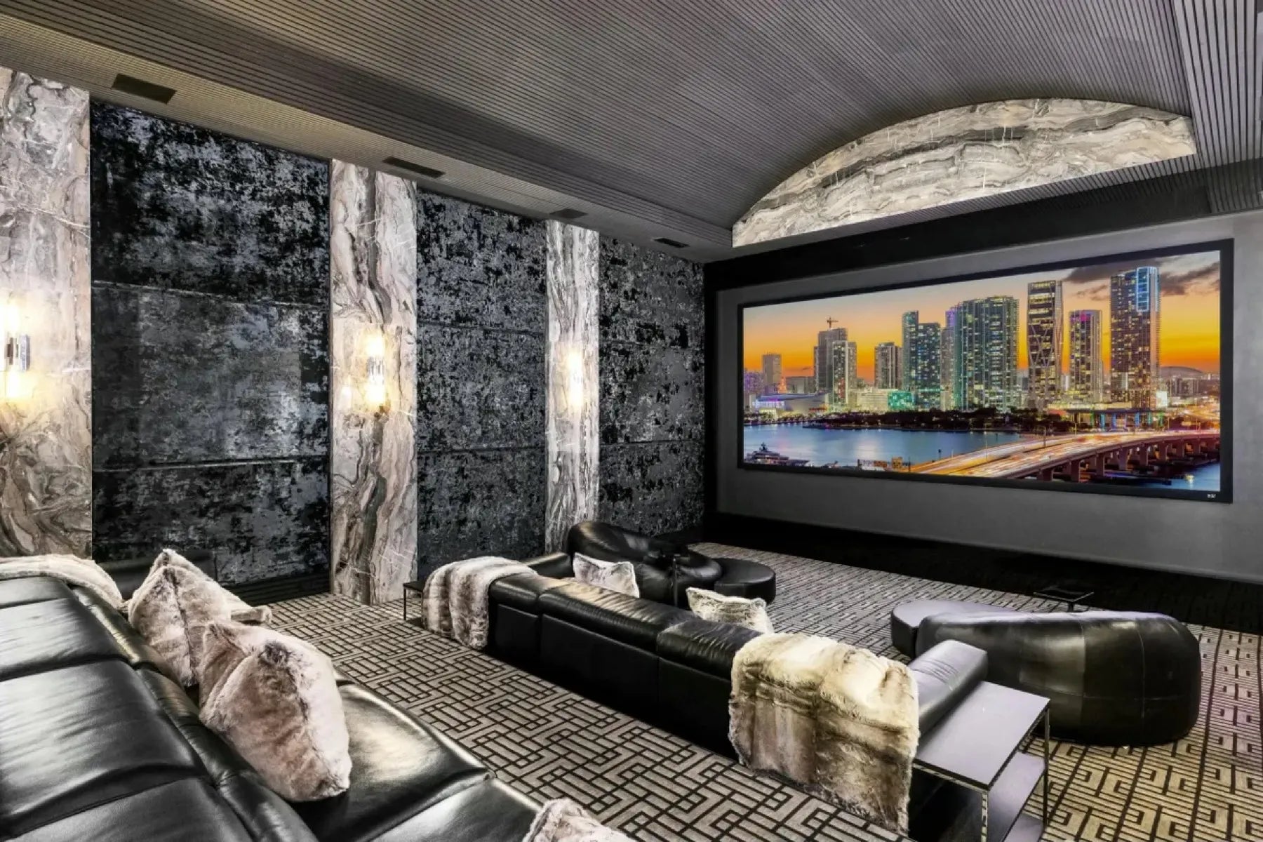 Home theater with black distressed upholstered wall panels between marble pilasters, lit by wall sconces, facing a wide cinema screen